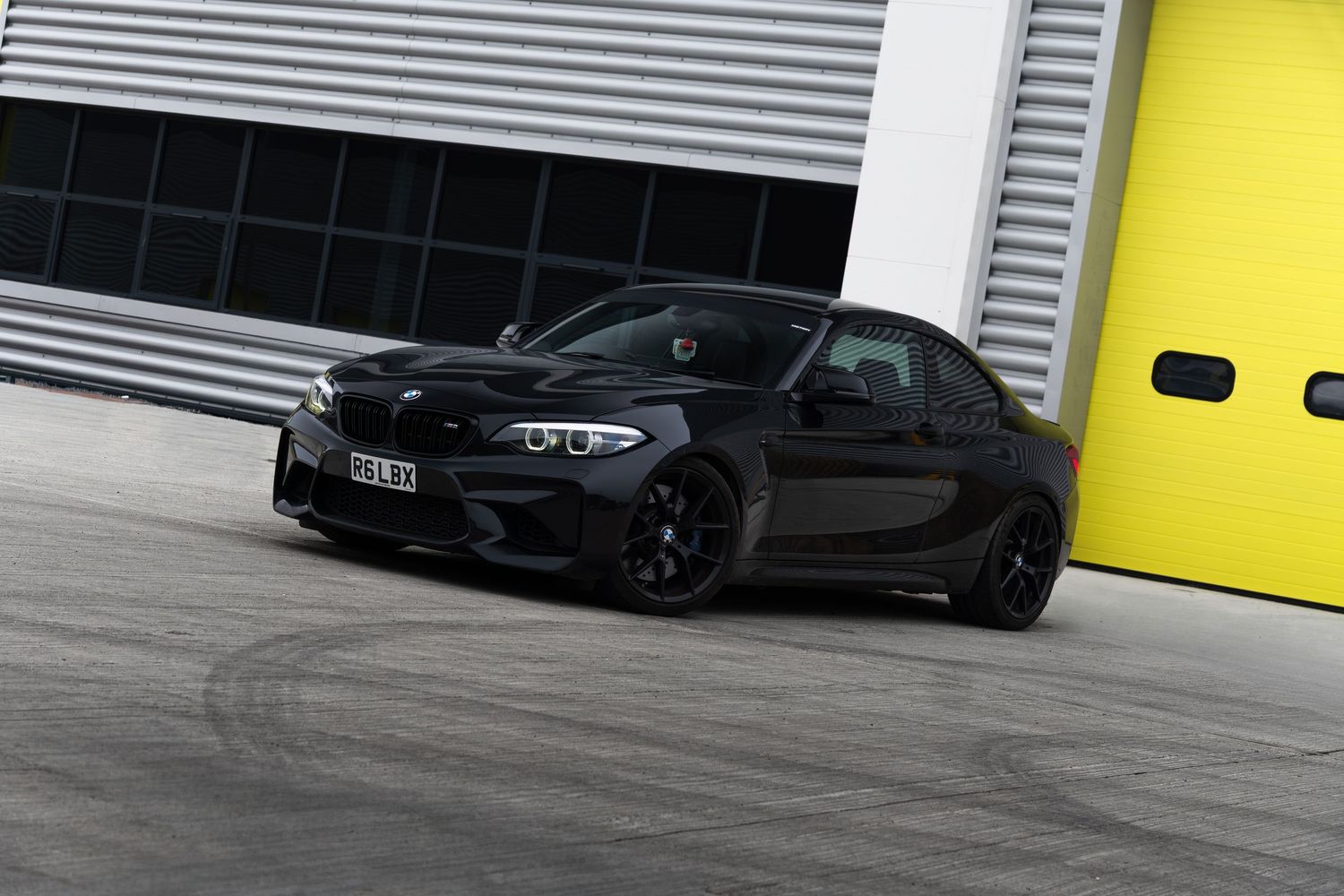 Black BMW M2 coupe driving in front of yellow garage doors on a wide concrete surface.