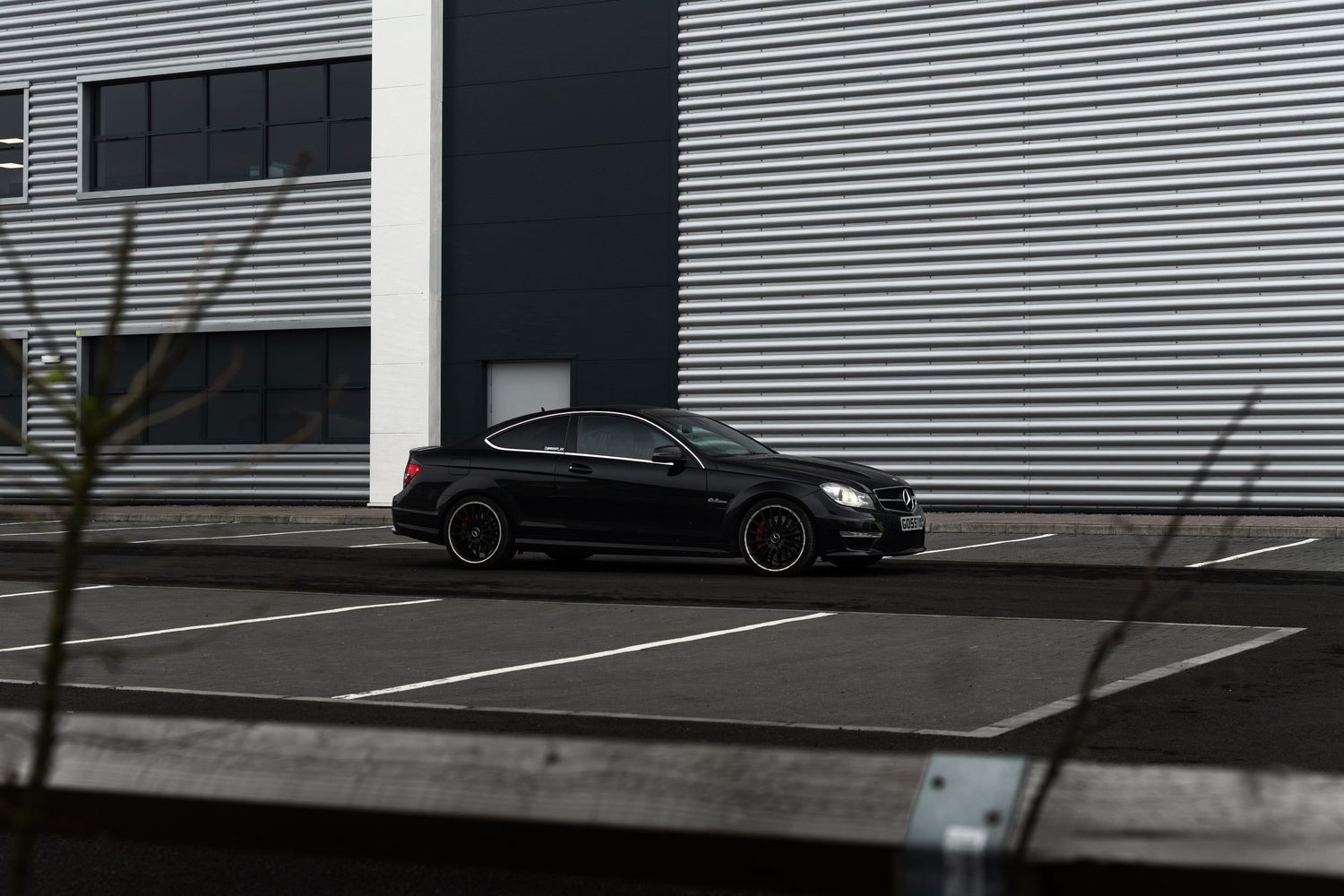 Matte black Mercedes-AMG C63 coupe parked in an empty lot beside a corrugated metal building.