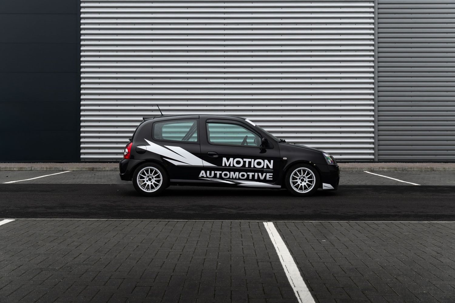 Full side profile of a black Motion Automotive Renault Clio on a dark tarmac surface against a metal wall.