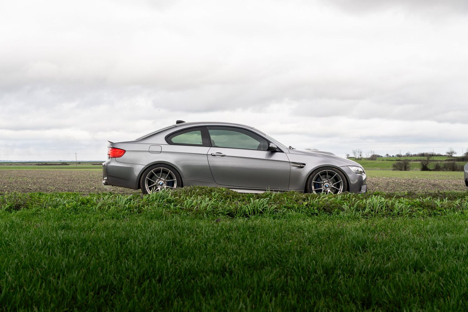 Side view of grey BMW M3 coupe with sport wheels parked on green countryside field.