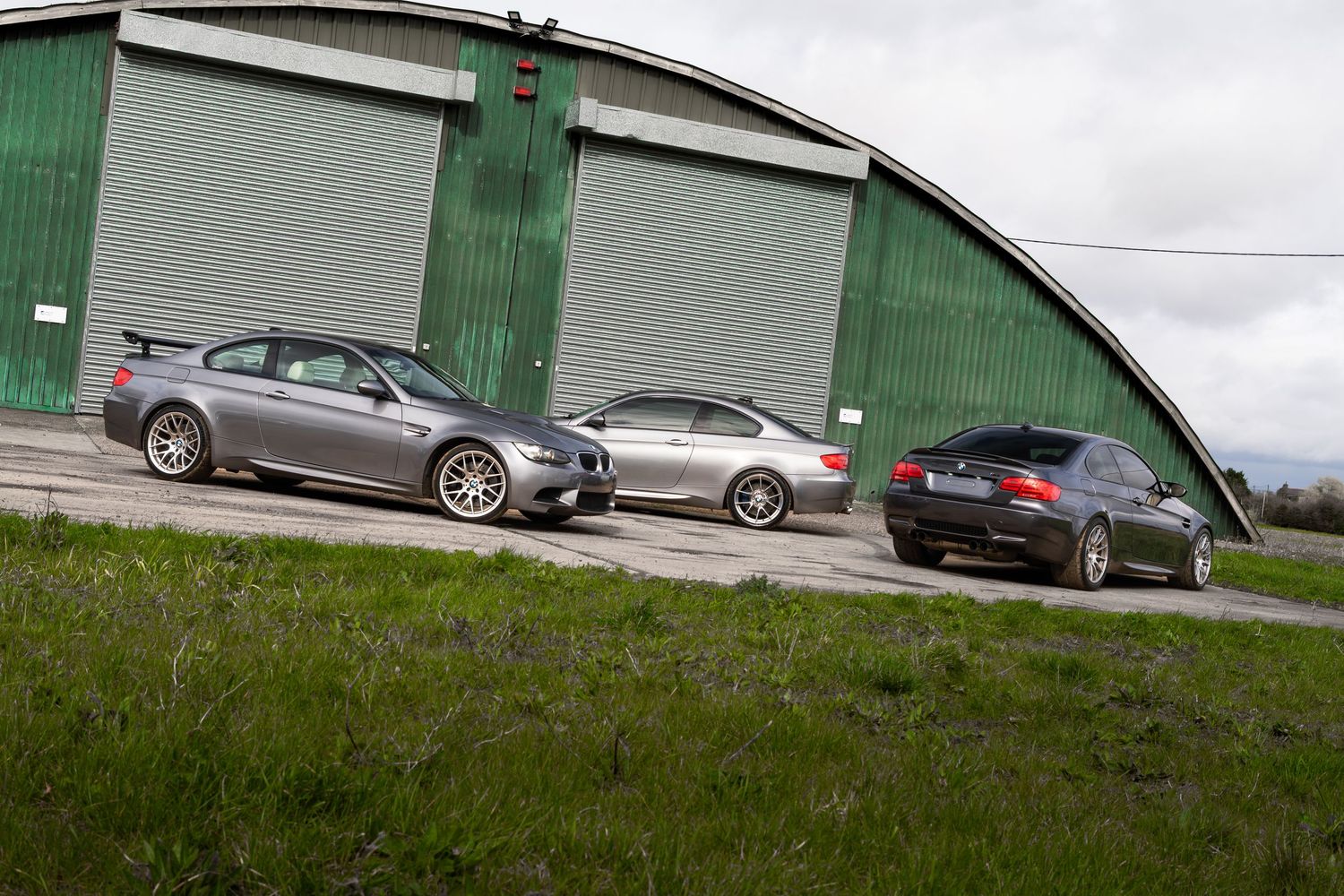 Three gray BMW M3 coupes lined up outside a large green arched metal hangar building.