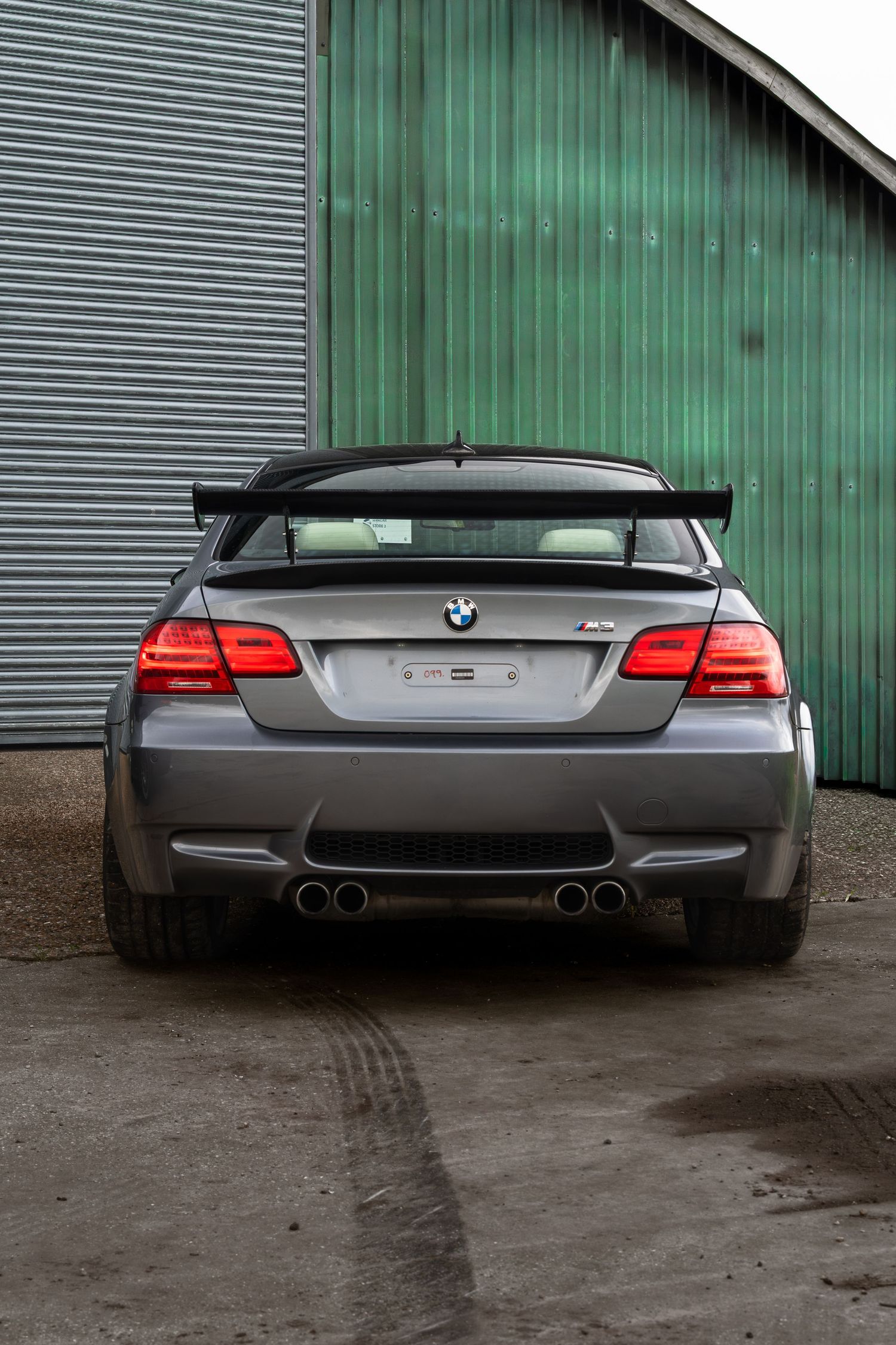 Rear view of a grey BMW M3 coupe with large spoiler and quad exhausts outside green hangar.