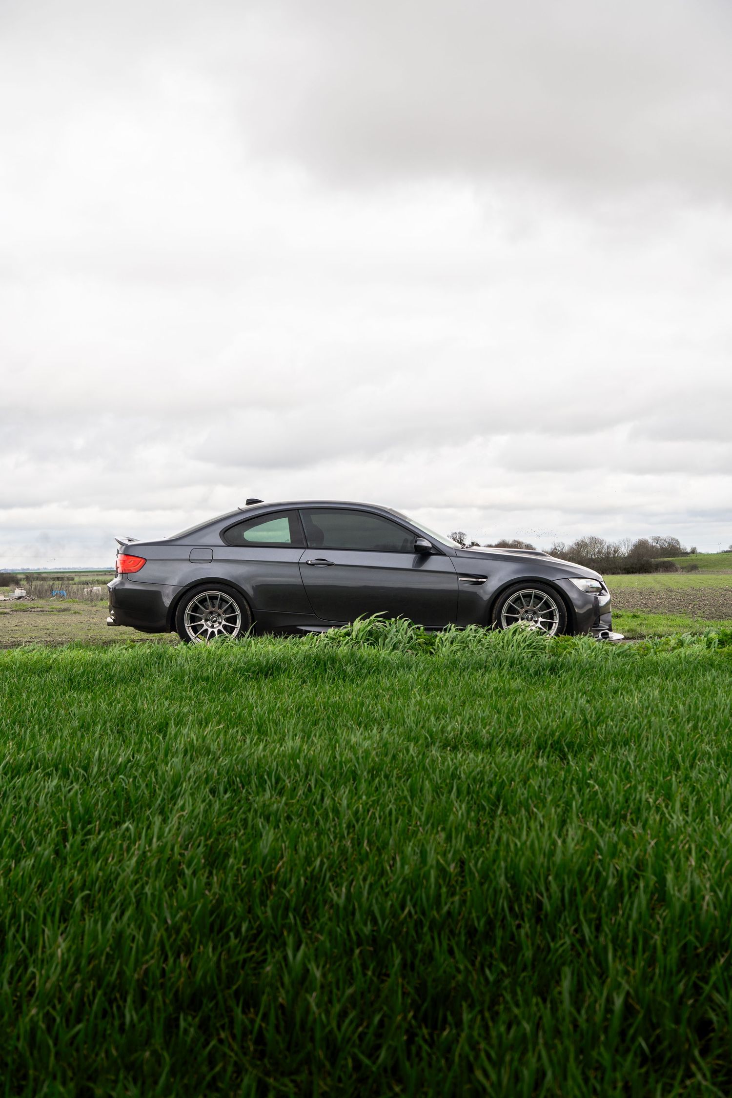 Dark grey BMW M3 coupe side view parked on vibrant green field under overcast sky.