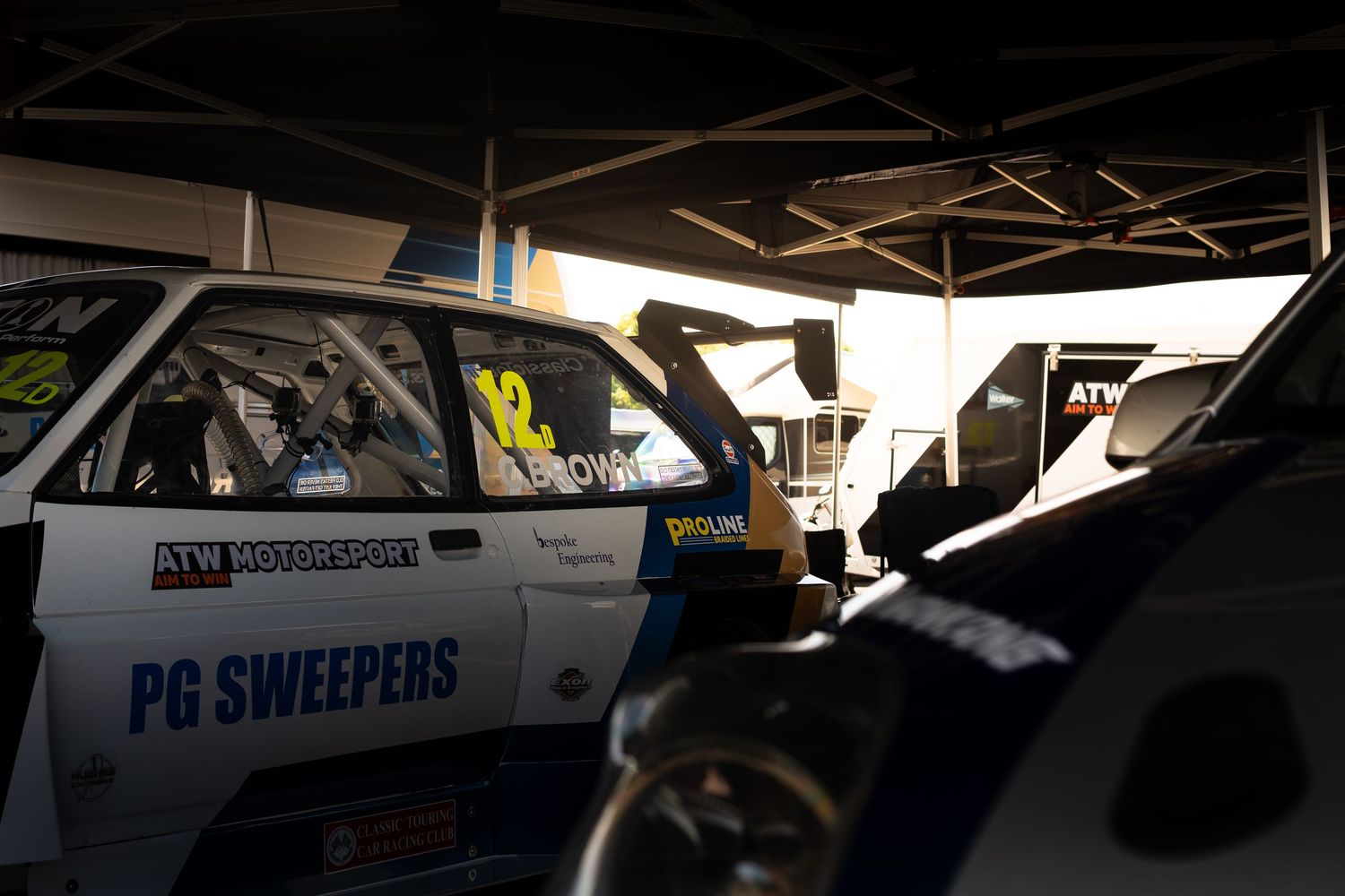 A white and blue PG Sweepers sponsored race car parked in a shaded pit garage.