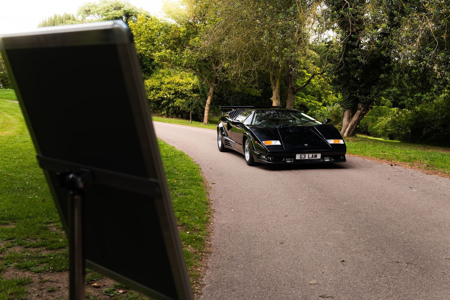 Black Lamborghini Diablo parked on a tree-lined driveway, captured from behind a photography reflector panel.