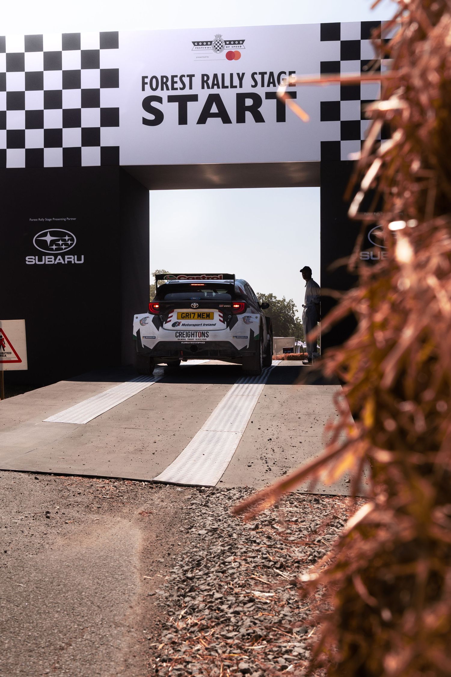A Subaru rally car at the Forest Rally Stage start line under a checkered flag arch.