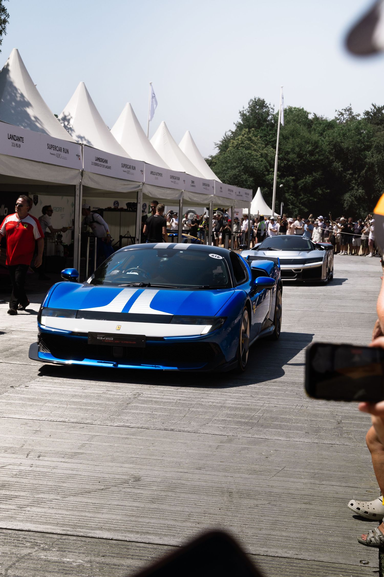Blue Ferrari with white racing stripes at an outdoor supercar event, surrounded by spectators and white tent pavilions.