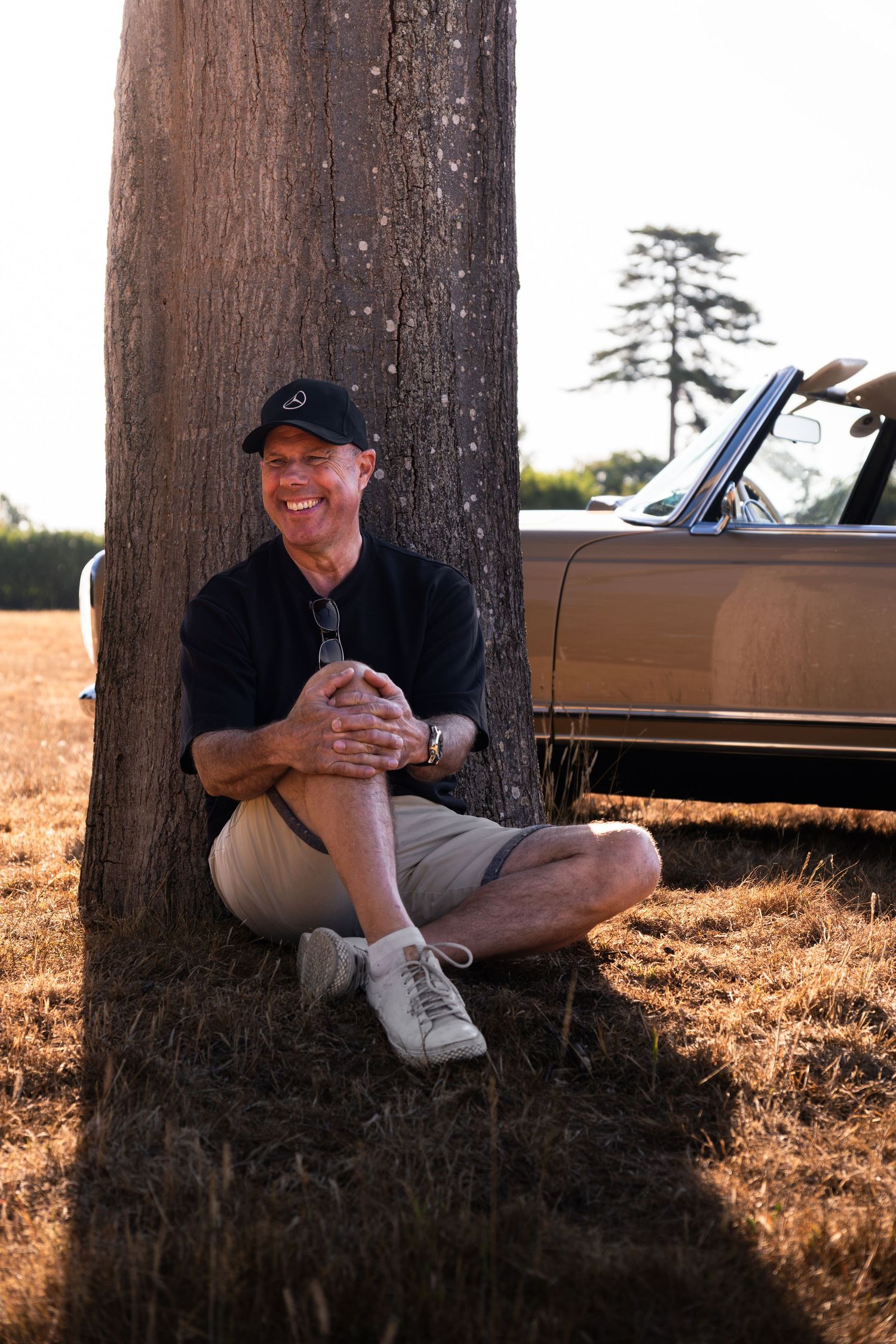 Man in black cap sits cross-legged against a tree beside a vintage convertible car in a sunny field.