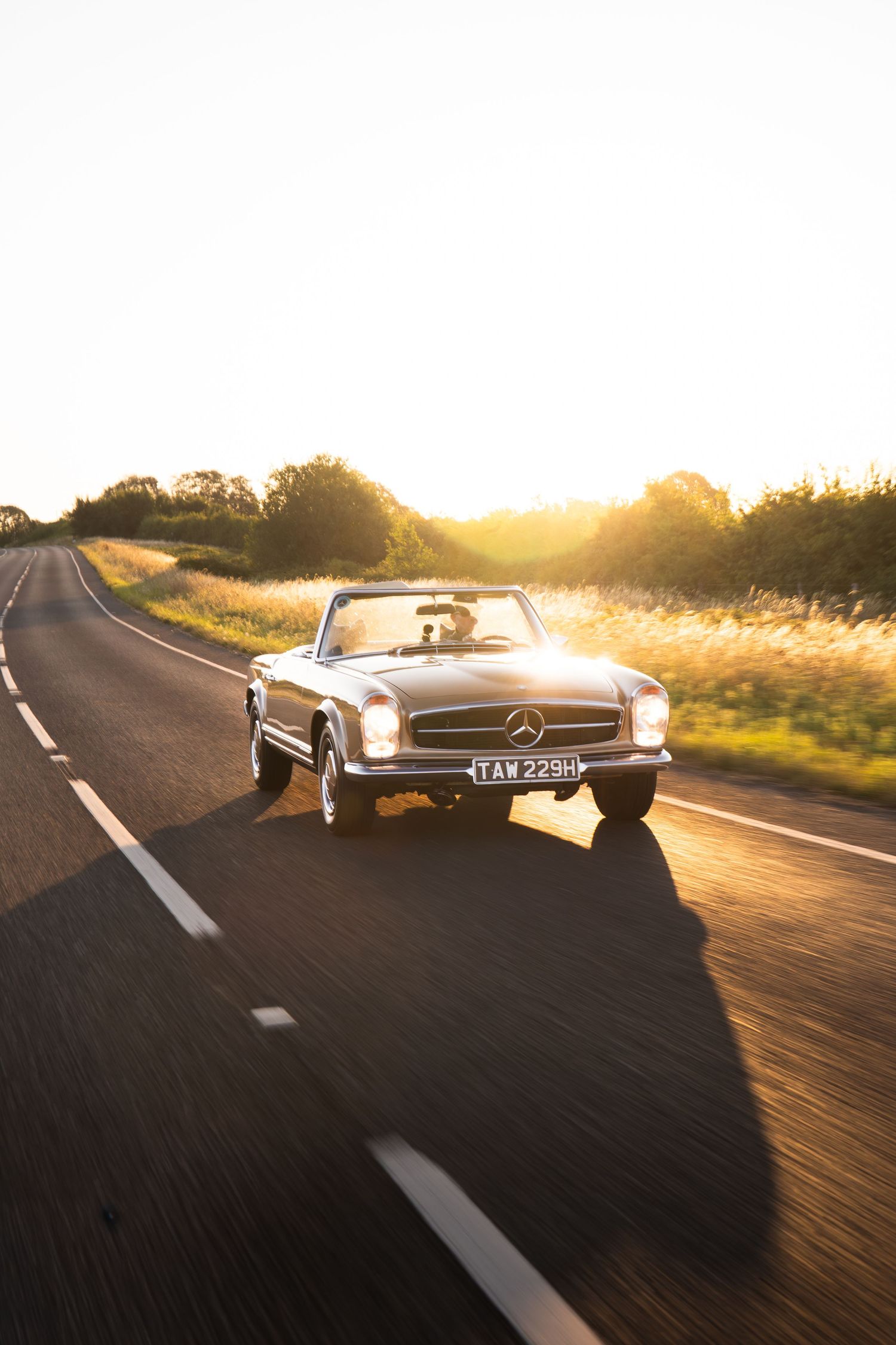 Classic Mercedes-Benz convertible driving on a rural road at golden sunset hour.