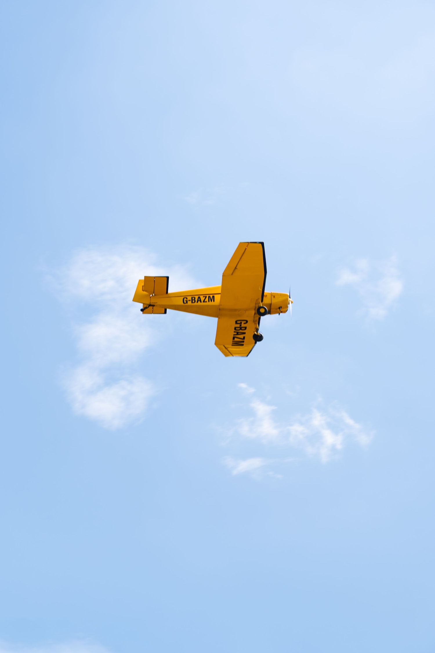 Yellow light aircraft G-BAZM flying overhead against a clear blue sky with light clouds.