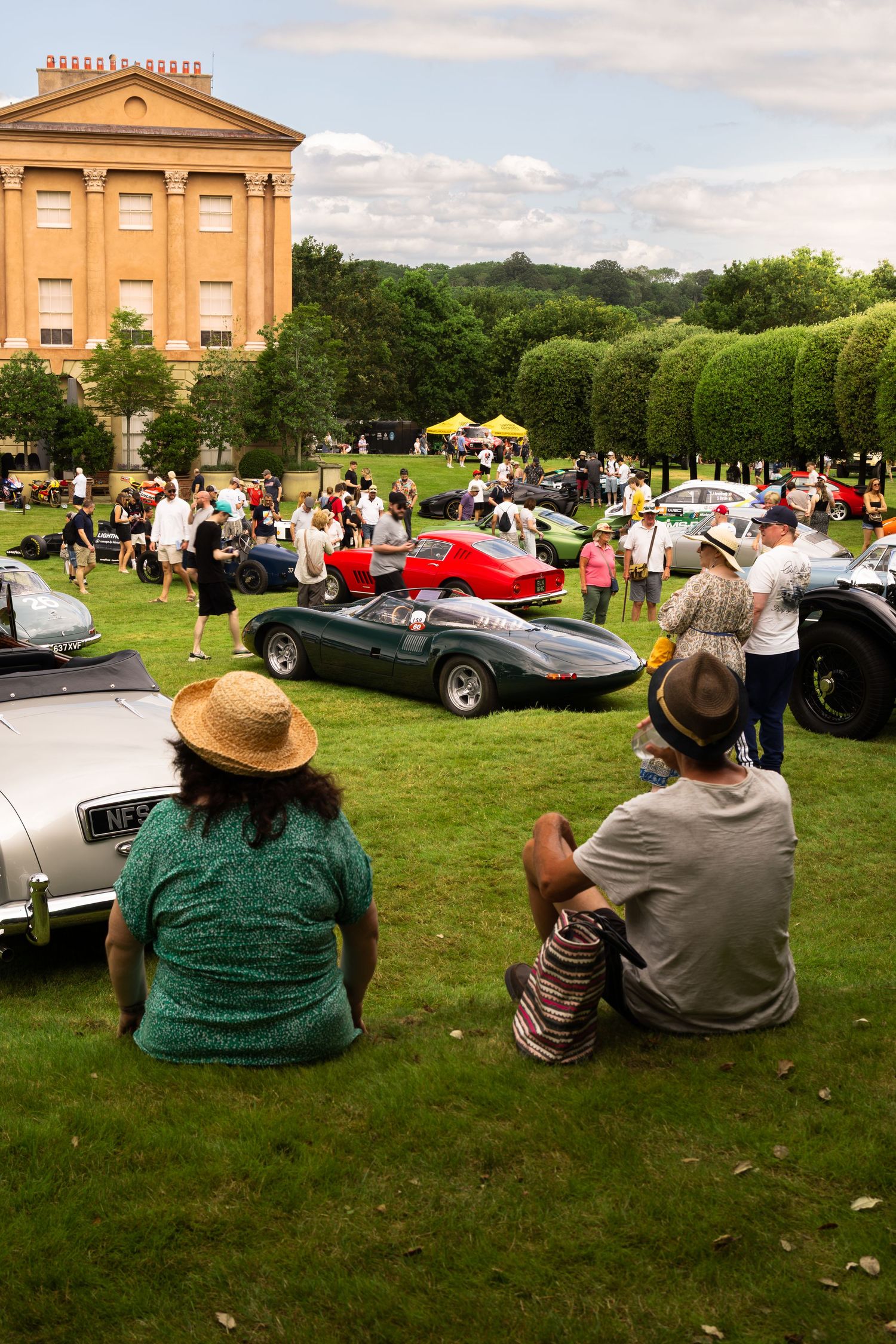 Spectators sit on grass watching classic cars at an outdoor vintage car show near a grand historic building.