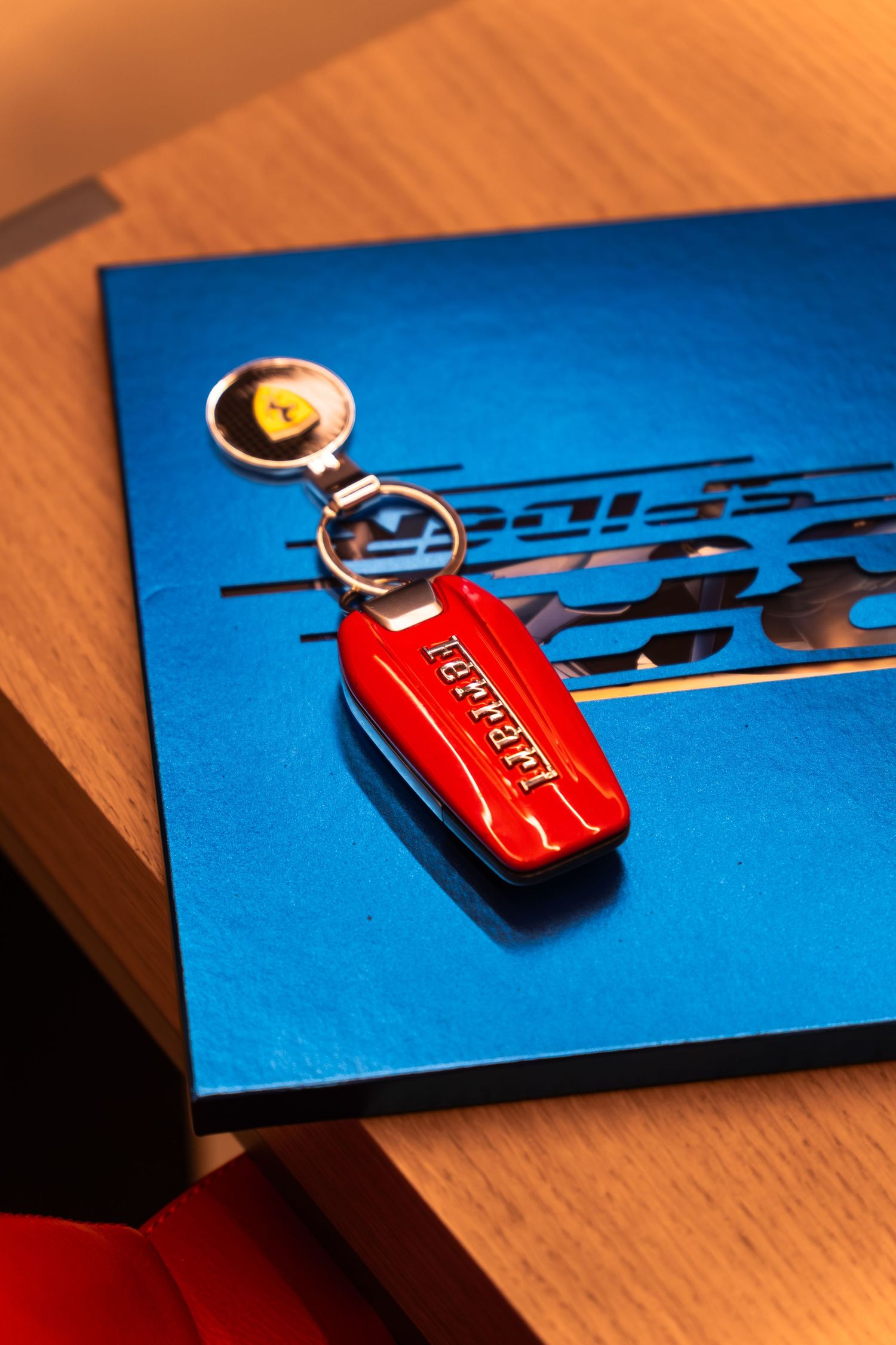 Red Ferrari branded keychain with prancing horse logo resting on a blue Ferrari book on a wooden surface.