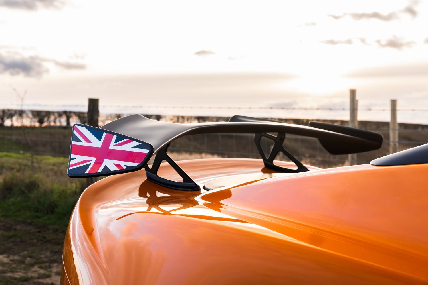 Close-up of orange Lotus Elise rear spoiler with Union Jack mirror detail against sunset sky.