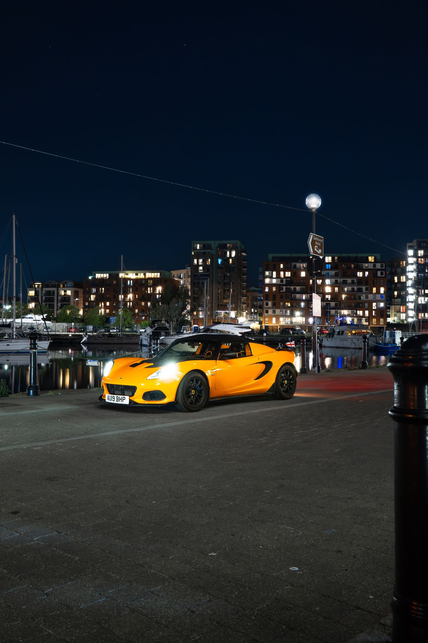 Bright yellow Lotus Elise at a waterfront marina at night with city buildings illuminated behind.
