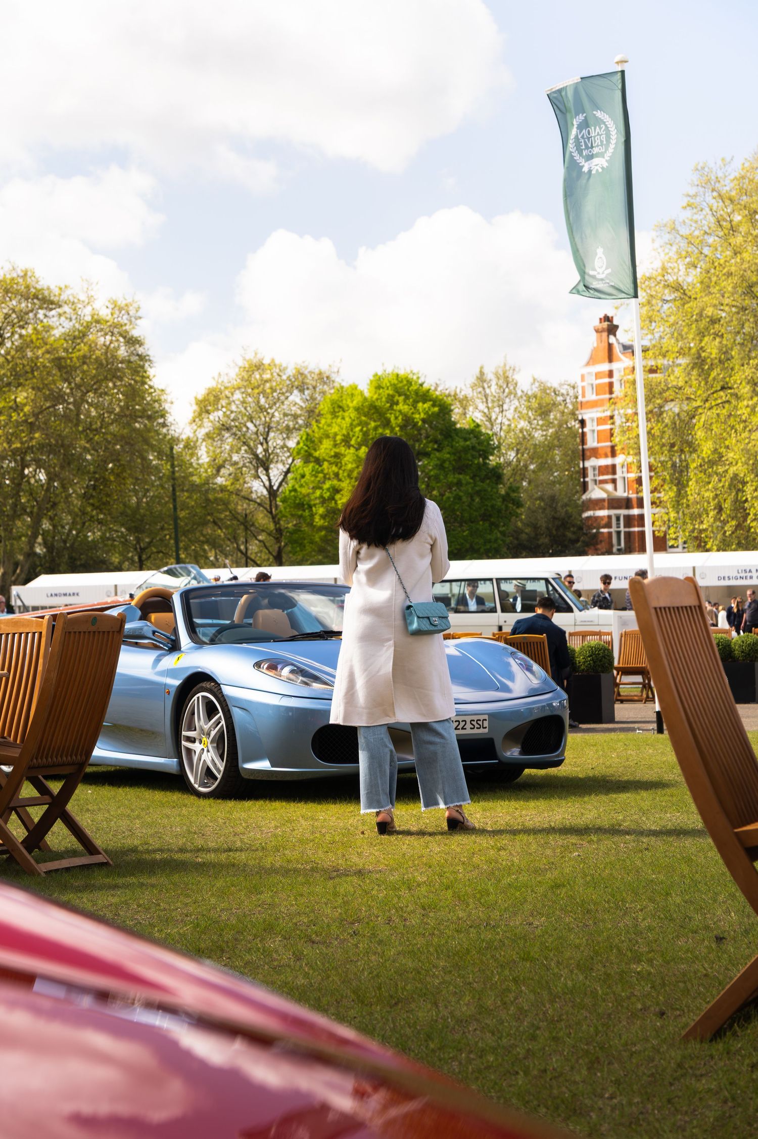 Woman in white coat admiring a silver-blue Ferrari F430 Spider at an outdoor car exhibition.