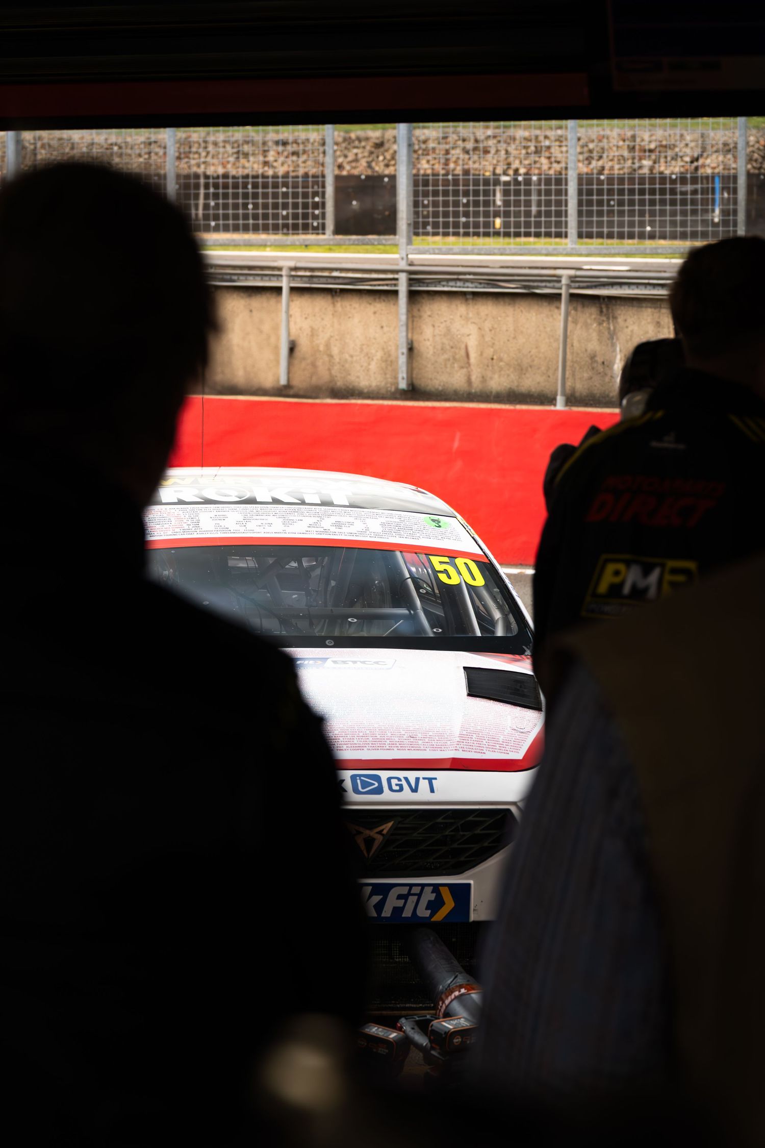 Silhouetted spectators watch a white race car numbered 50 in the pit lane at a motorsport event.