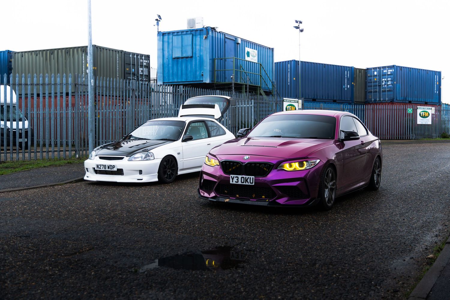 A white Honda Civic and purple BMW M2 parked in front of shipping containers.