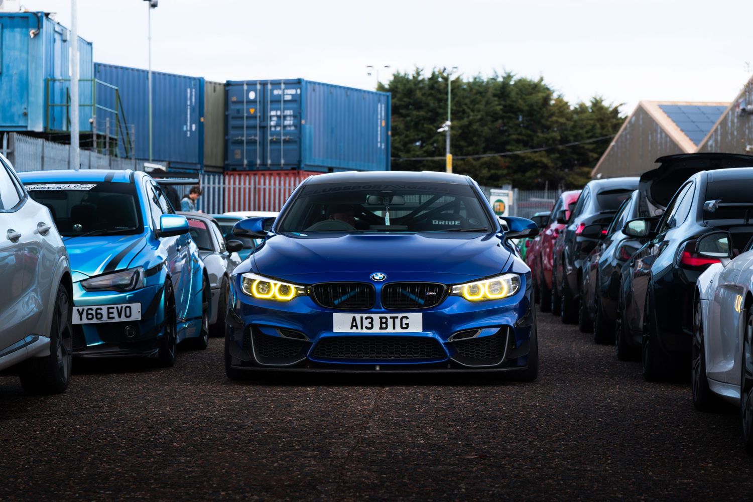 A blue BMW M4 with yellow angel eyes parked between rows of cars at a car meet.