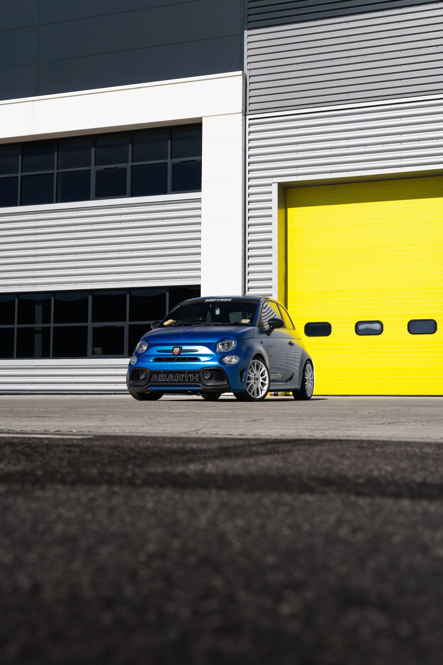 Blue Abarth 500 parked in front of yellow garage doors at a motorsport facility.