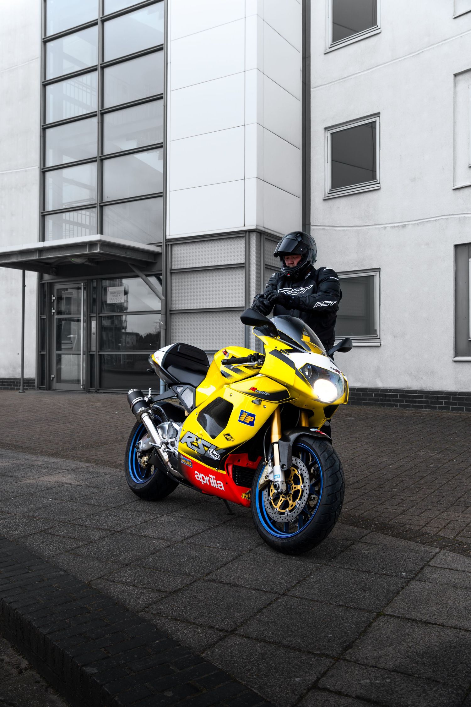 Rider in black gear sitting on a yellow Aprilia RSV sport motorcycle outside an office building.