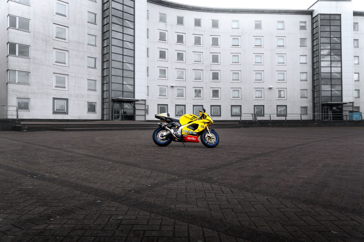 Yellow Aprilia RSV motorcycle centered on wide cobblestone plaza in front of a curved white building.
