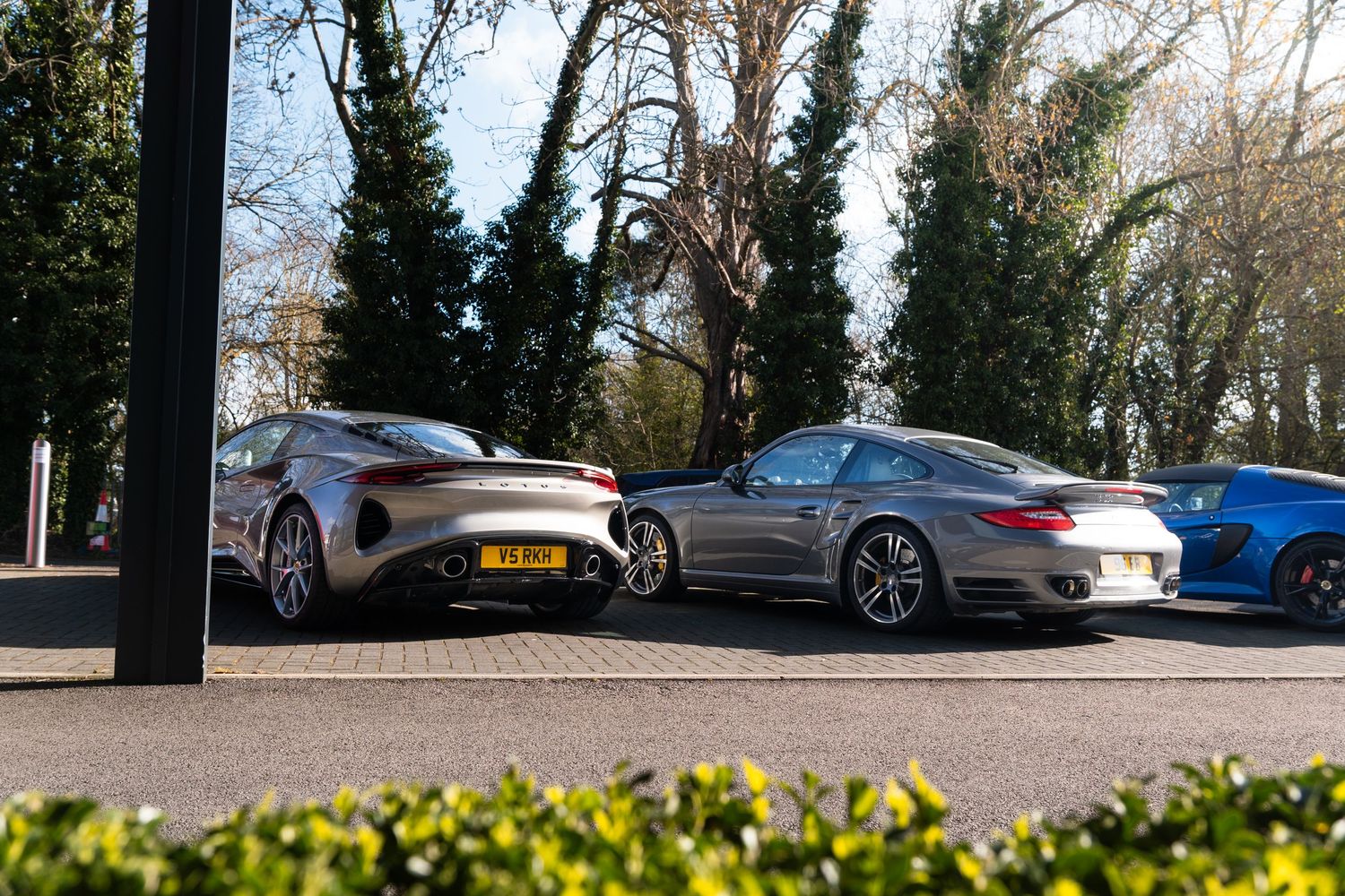 Three luxury sports cars including an Aston Martin and Porsche 911 Turbo parked in a sunlit lot surrounded by trees.