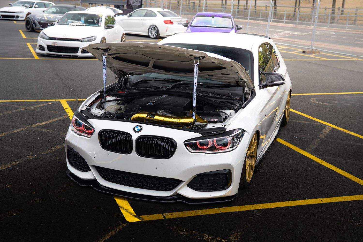 White BMW with open hood revealing yellow engine bay modifications in an outdoor parking lot.