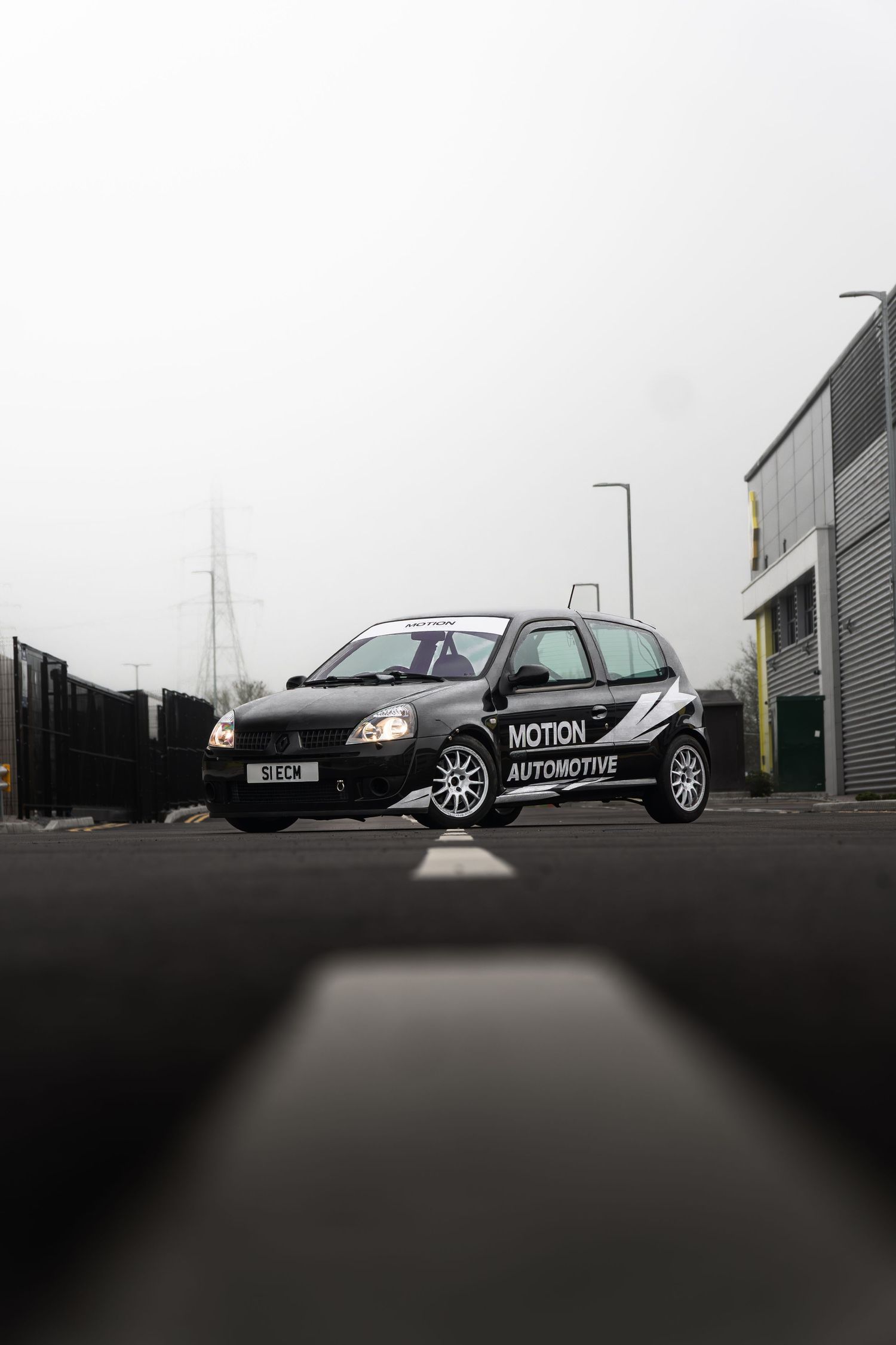 Low angle shot of a black Motion Automotive Renault Clio on a wet road with industrial buildings in foggy background.