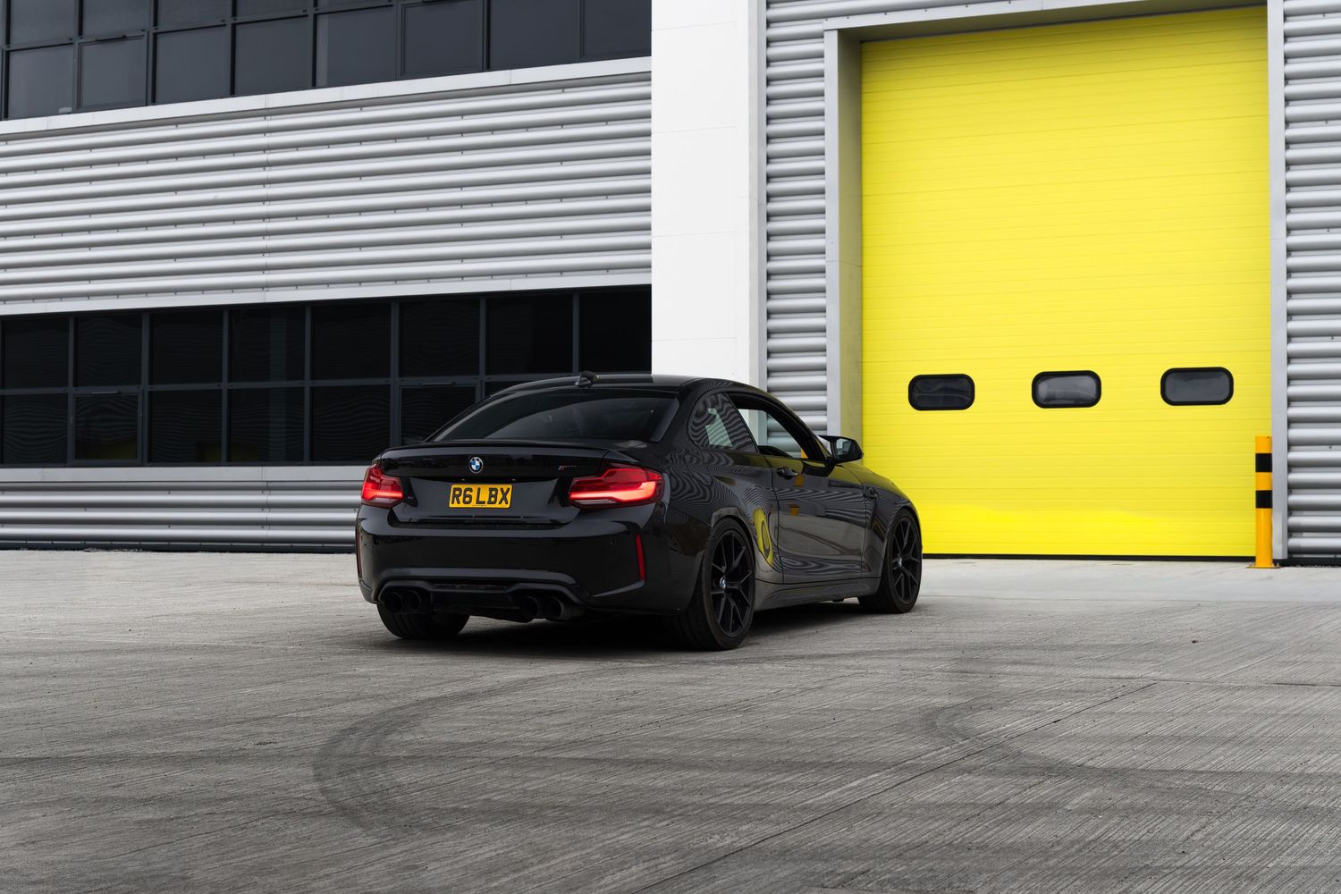 Three-quarter rear view of a black BMW M2 coupe parked outside yellow industrial doors.