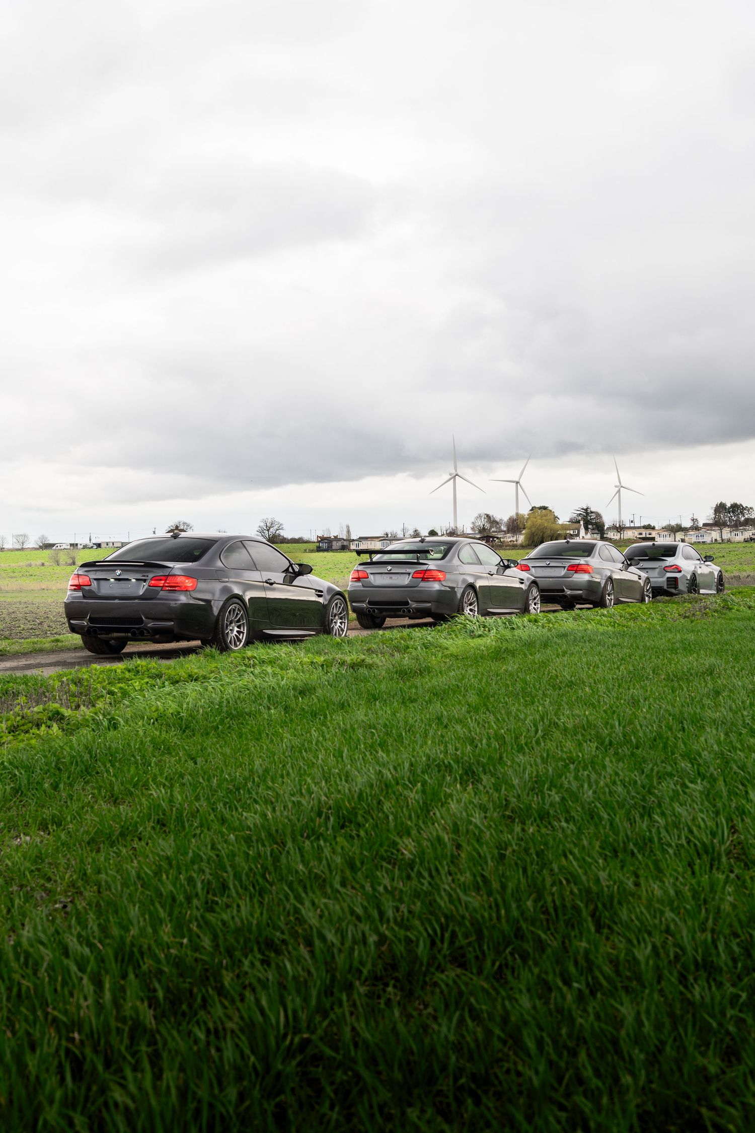 Rear view of several BMW M3 coupes parked on rural grass field near wind turbines.