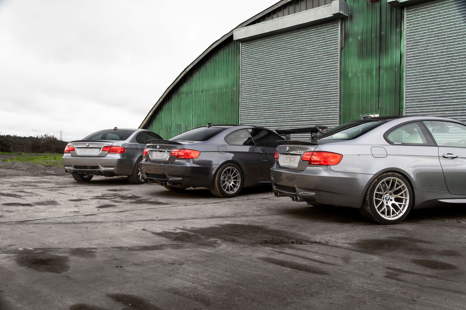 Three BMW M3 coupes photographed from the side in a wet parking area beside a green hangar.