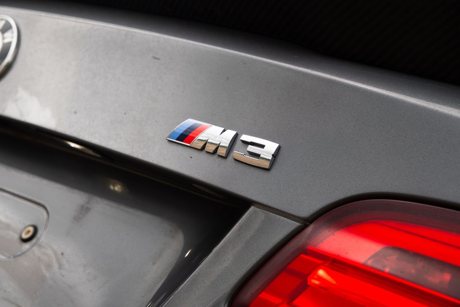Close-up of BMW M3 trunk badge with red brake light visible on gray painted bodywork.
