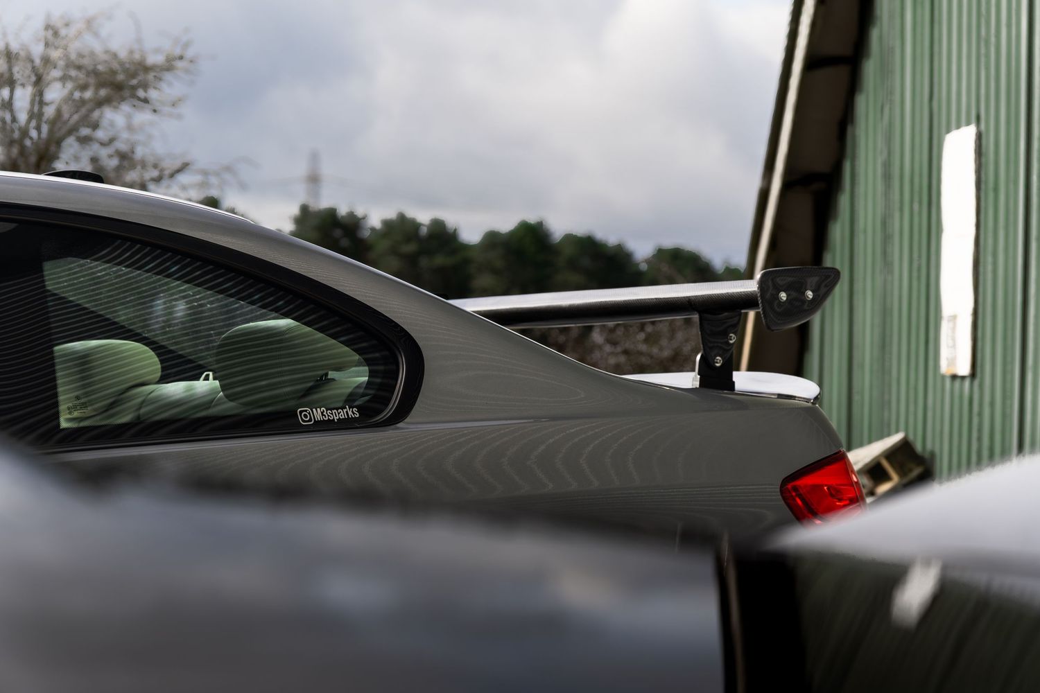 Close-up of a grey BMW M3 coupe rear spoiler and roofline against green building.