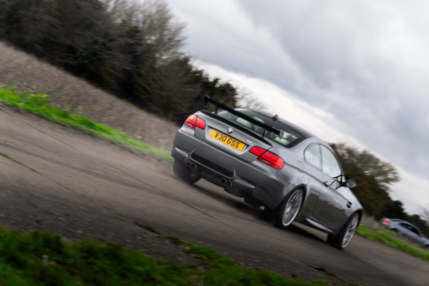 Rear view of a silver BMW M3 E92 coupe accelerating on a rural road.
