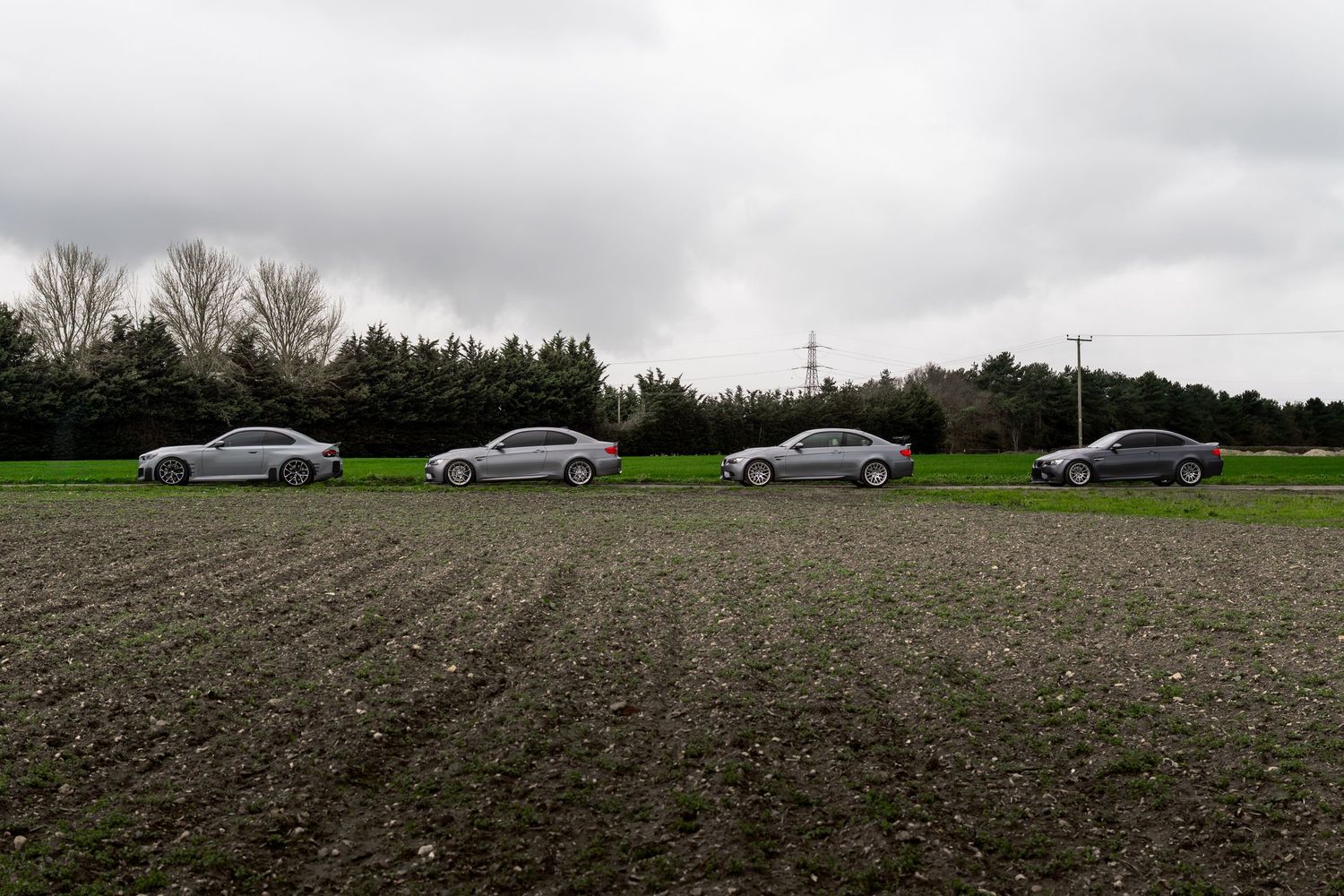 Four BMW M3 coupes parked in a row on a plowed field with trees and a pylon in background.