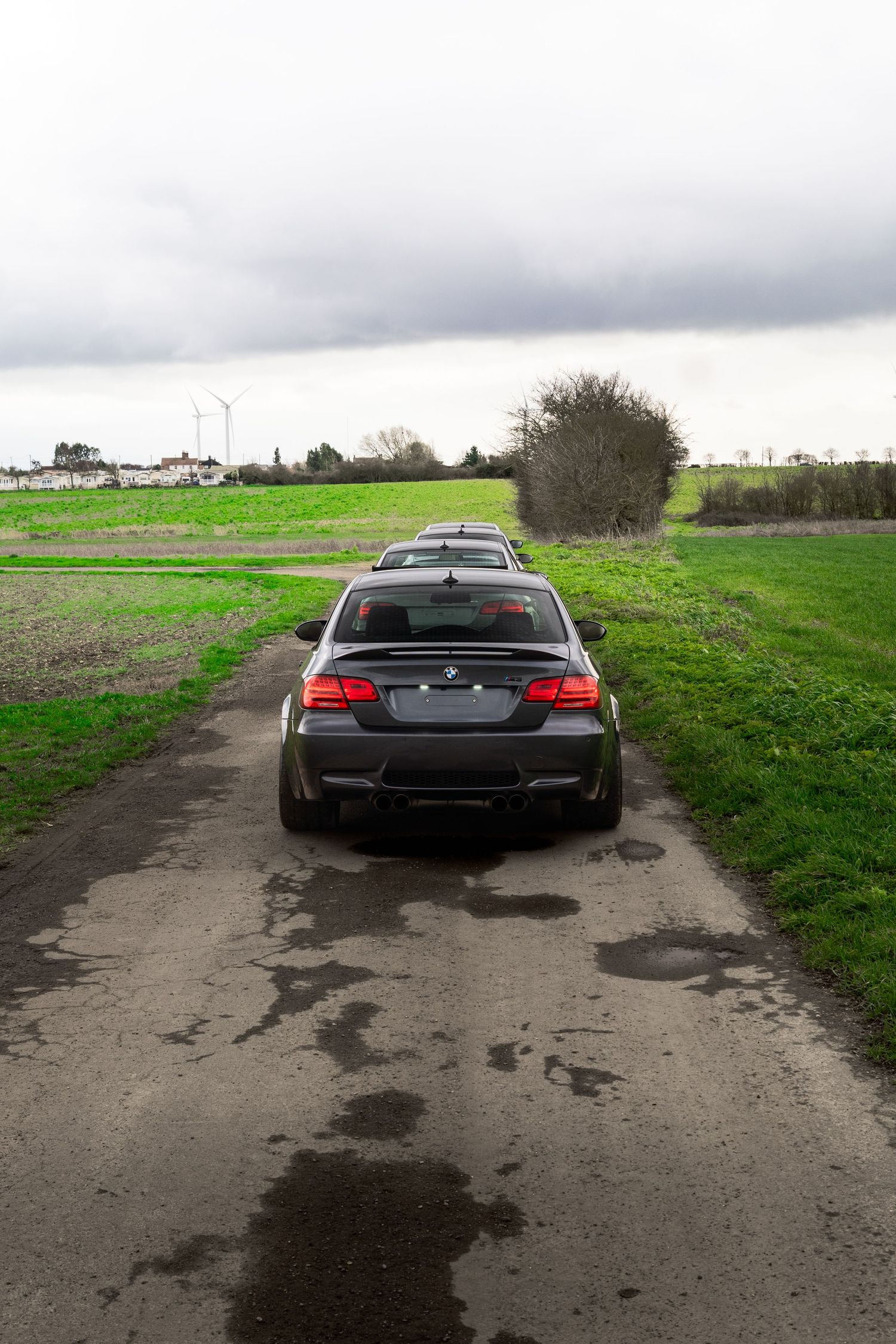 Dark gray BMW M3 coupe viewed from behind on a narrow rural road with green fields.