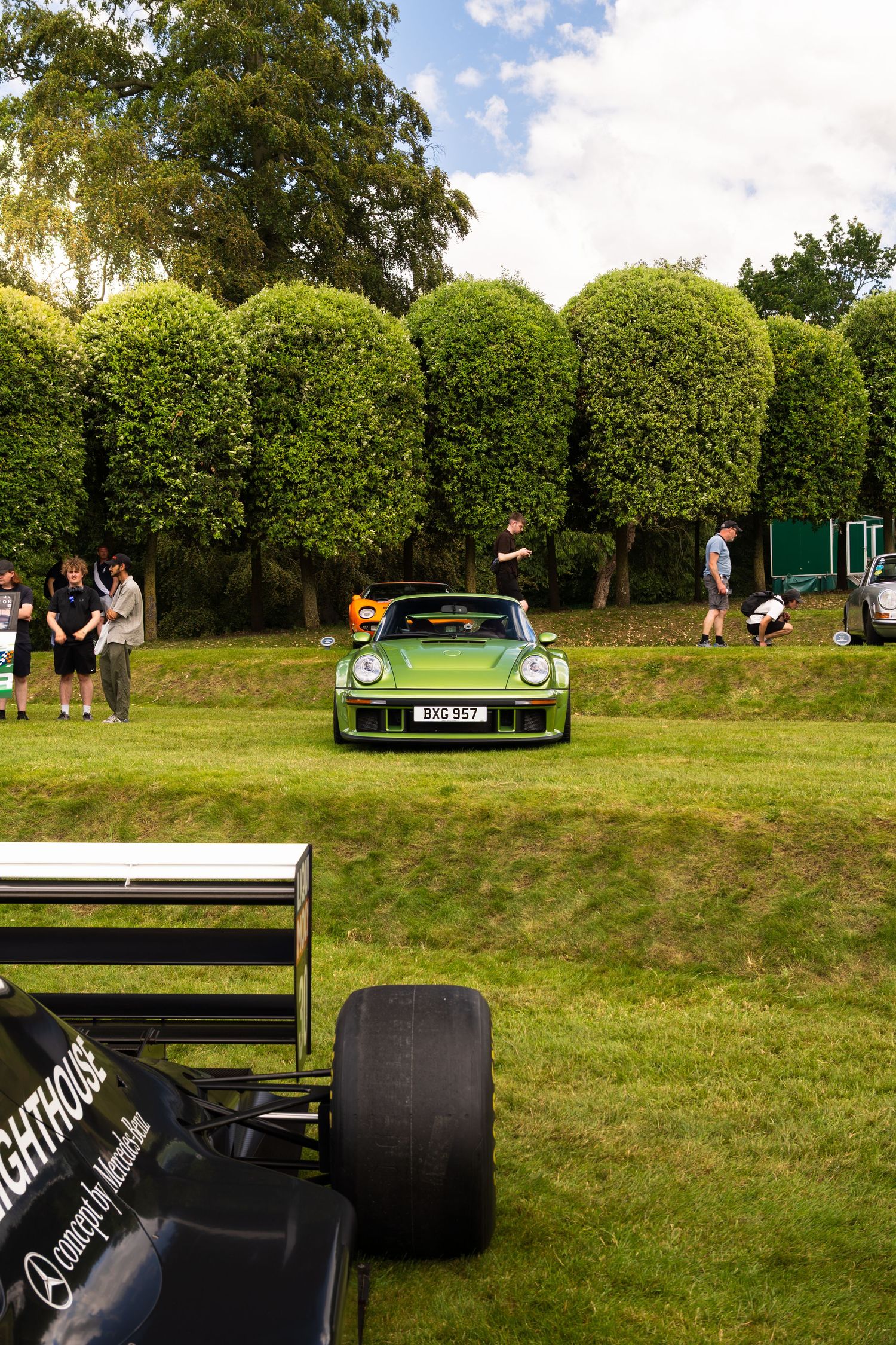 Green Porsche 911 on display at an outdoor classic car show, with manicured hedges and spectators in the background.