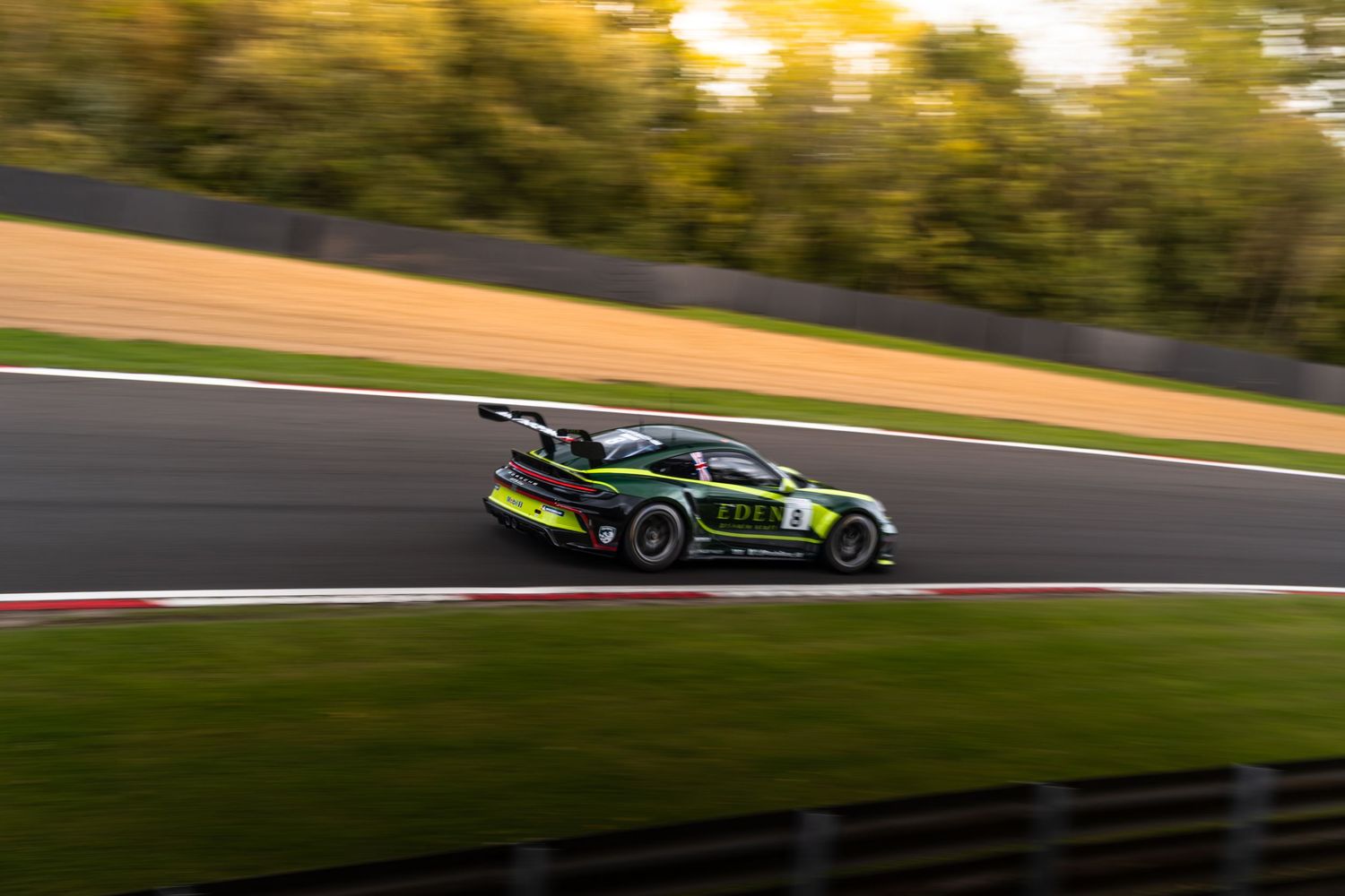 Green Porsche GT3 race car captured mid-corner on a racing circuit surrounded by autumn trees.