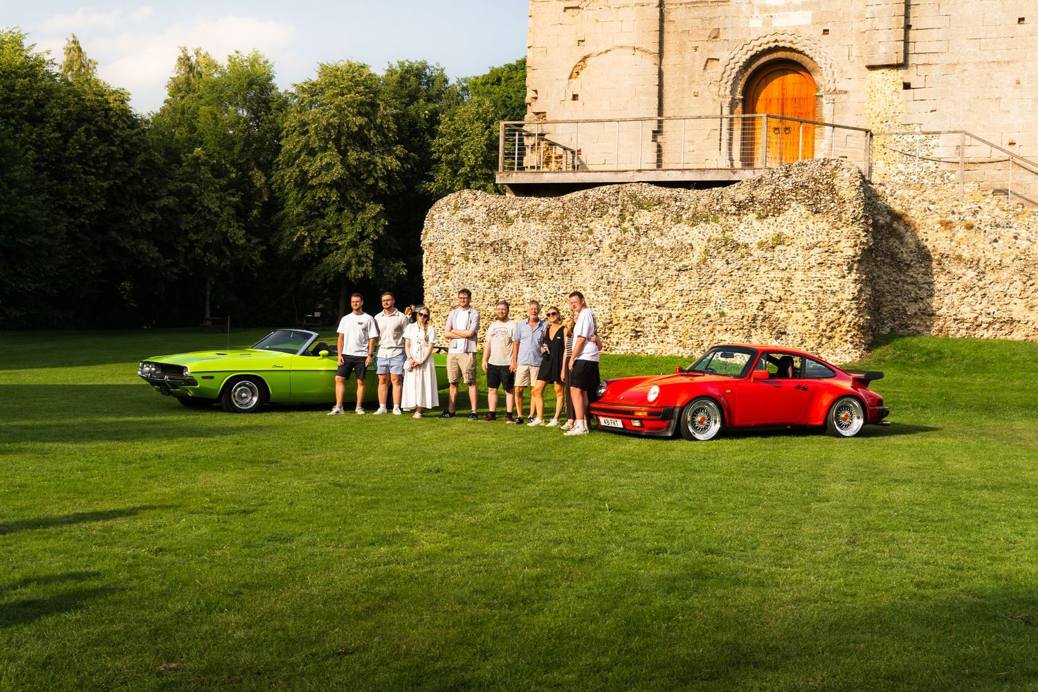 Group of people posing between a green classic Dodge Challenger and red Porsche 930 Turbo on a lawn by a stone building.
