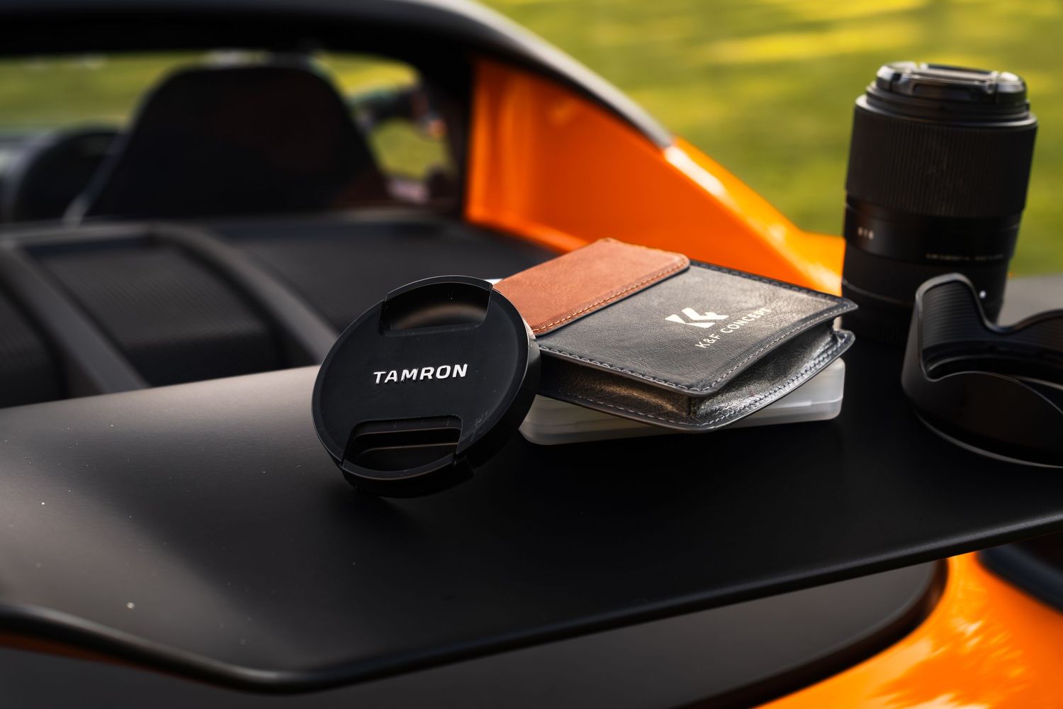 Tamron lens cap and camera lens on orange car dashboard with brown wallet outdoors.