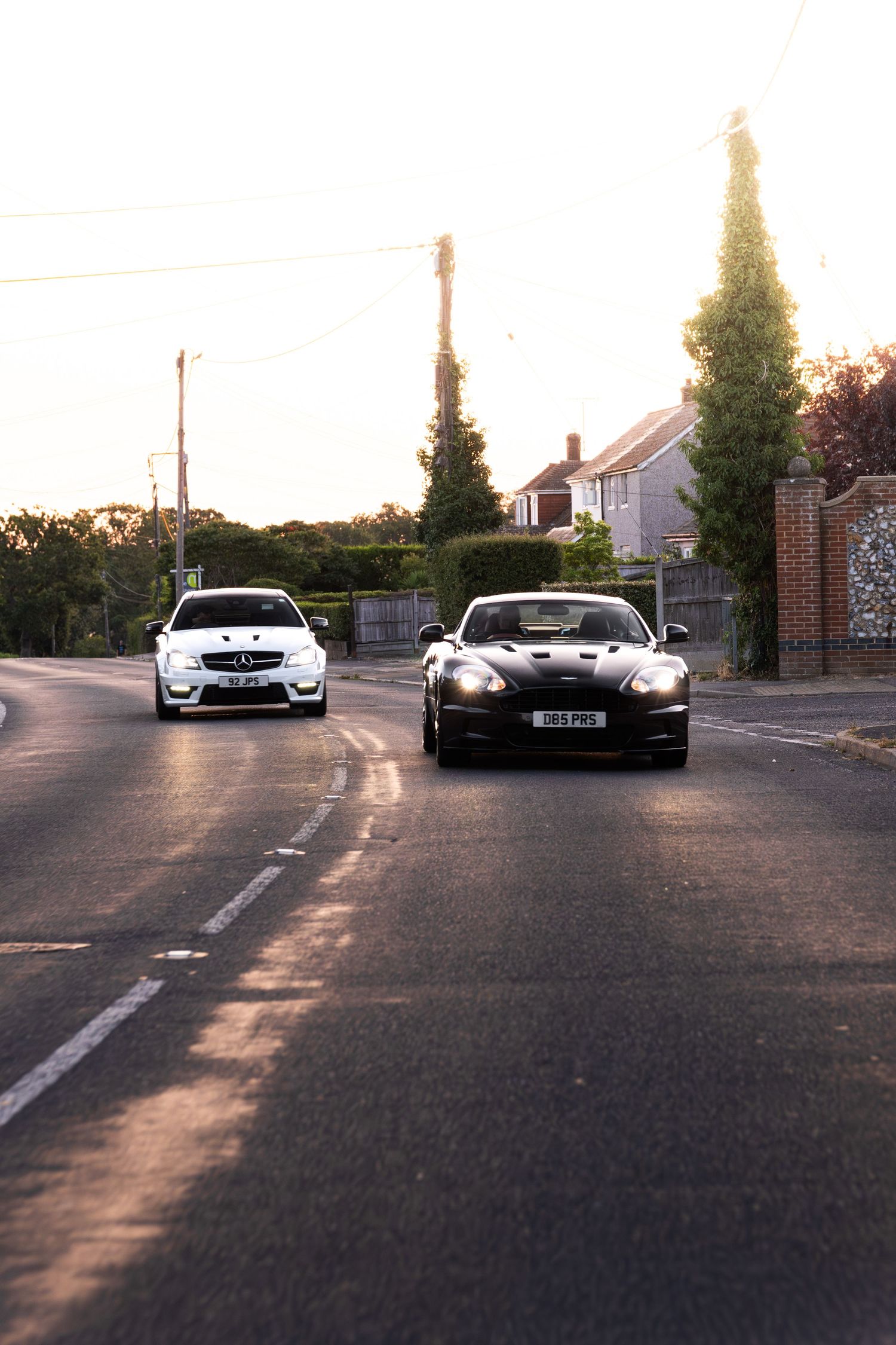 Black Aston Martin and white Mercedes driving on a quiet suburban road at golden hour sunset.