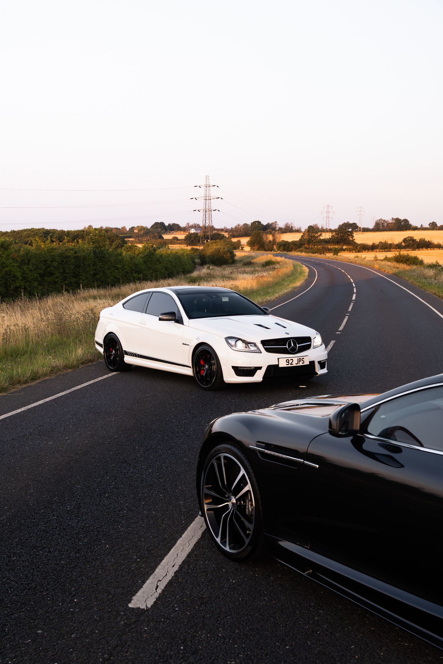 Two Mercedes-AMG C63 coupes, one white and one black, parked on a winding rural road at dusk.