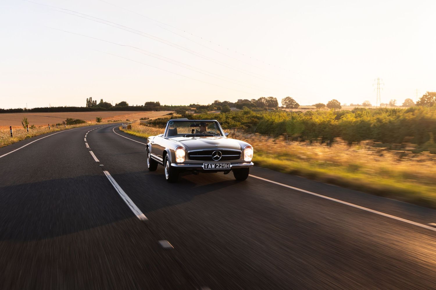 A classic Mercedes-Benz convertible drives along a scenic country road at golden hour.