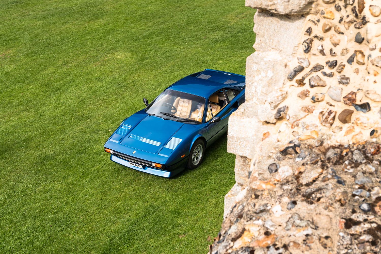 Blue Ferrari 308 GT4 sports car parked on green grass beside a stone wall, aerial view.