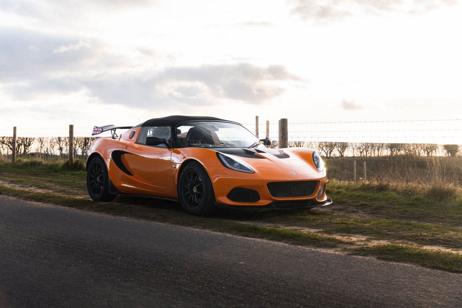 Orange Lotus Elise sports car parked on rural roadside under cloudy sky.
