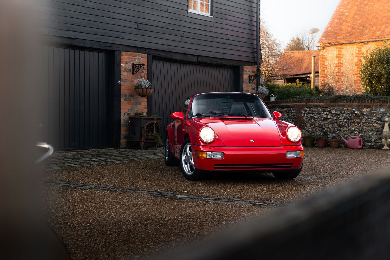 Classic red Porsche 911 with headlights on parked in front of dark garage doors at golden hour.