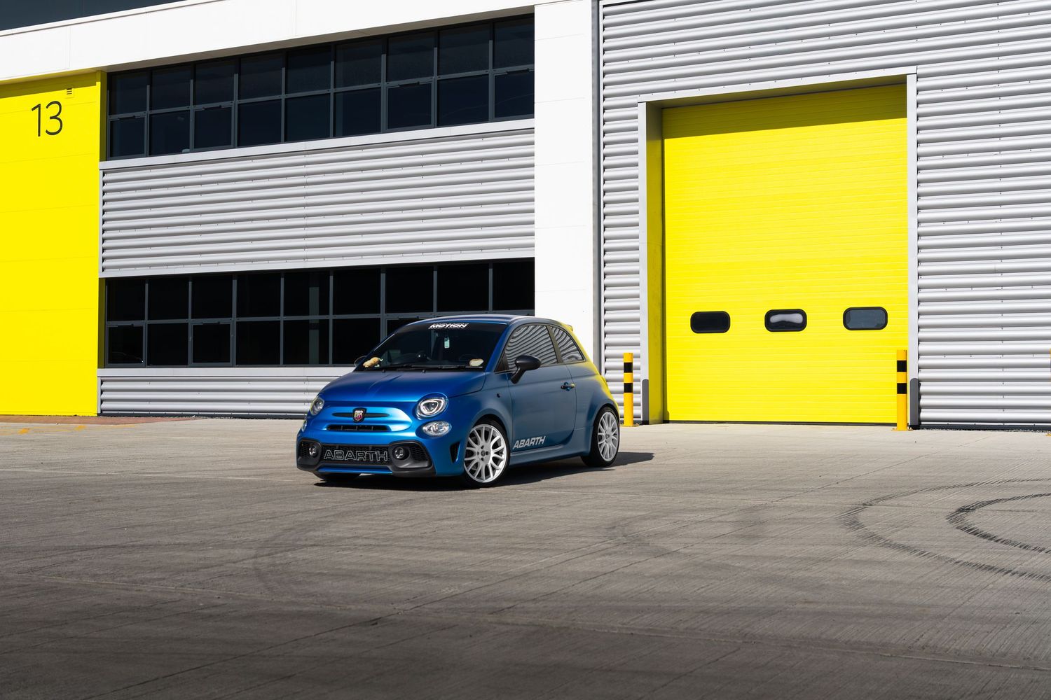 Blue Abarth 500 parked outside numbered industrial unit with yellow garage doors.