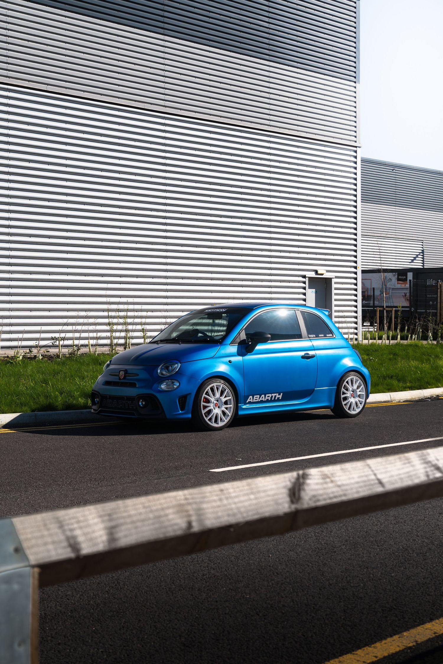 Blue Abarth 595 with white alloy wheels parked beside corrugated metal industrial building.