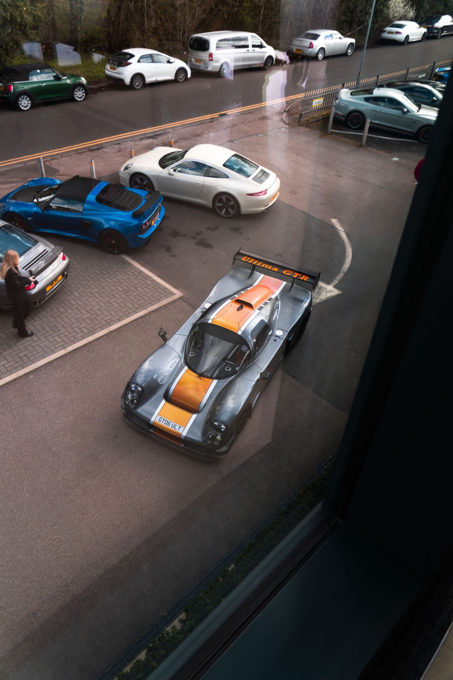 Overhead view of Ultima GTR supercar with orange stripes in wet car park alongside Porsche 911.