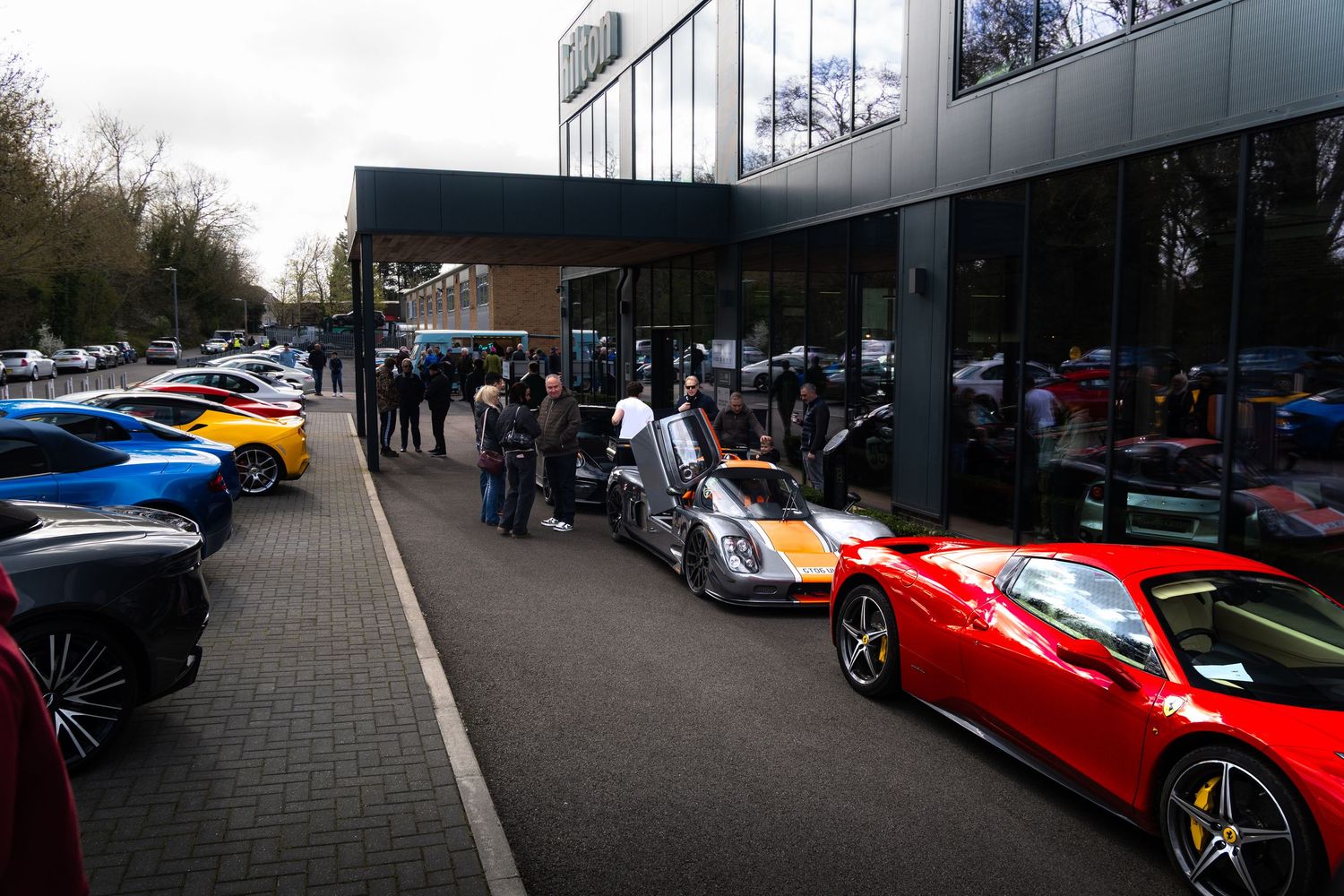 Collection of colorful supercars lined up outside glass-fronted dealership with crowd of enthusiasts.
